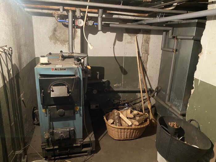 Gamla pannan i källare med rörledningar, korg med vedträn och sopkvast. Stenvägg med flagande färg i bakgrunden.