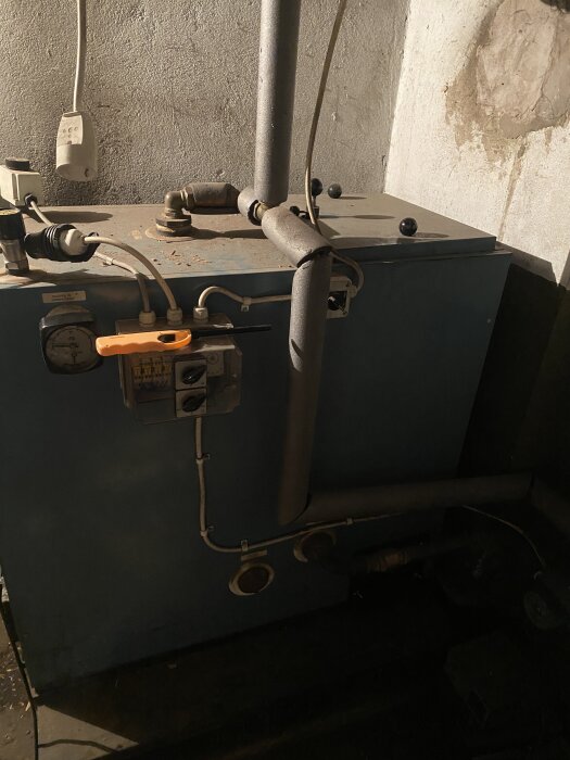 Gammal oljepanna med metallrör och kopplingsbox i ett mörkt pannrum, med smuts och rostiga ytor.