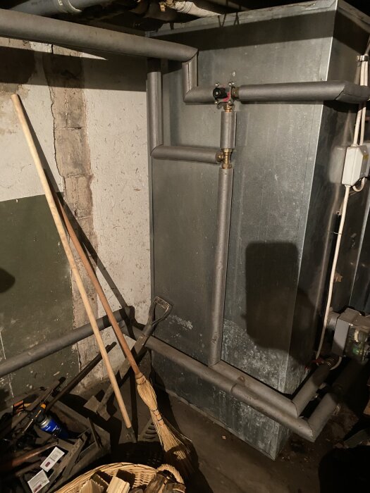 Källare med kopparrör och en stor värmepanna, omgiven av trädgårdsredskap som skyffel och kvast samt en korg med vedträn.