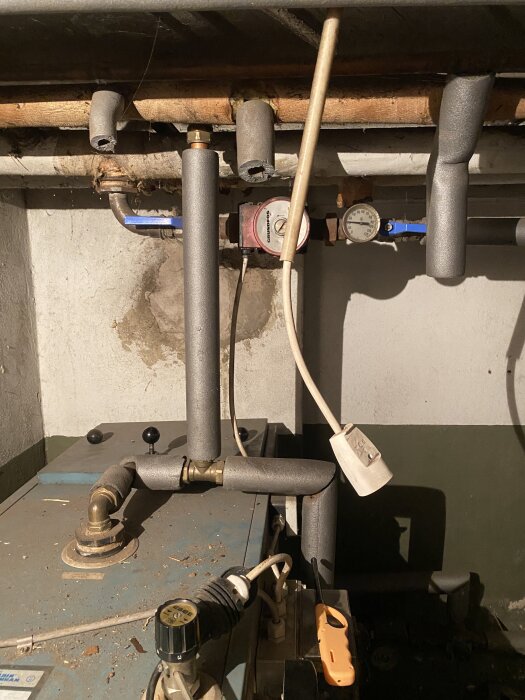 Rör och mätare i en källare, del av ett värmesystem. Några av rören är isolerade och det finns en pump och en termometer.