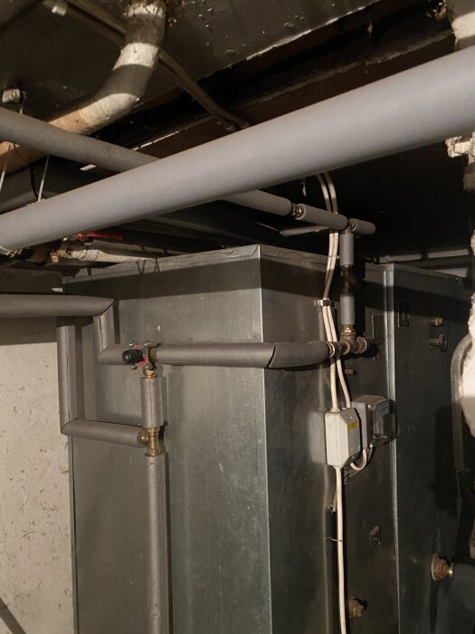 Pipes and wiring in a basement with insulation and a metallic unit, possibly part of a heating or plumbing system.