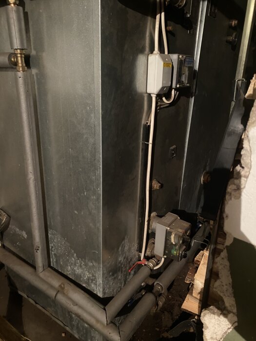 Gammal värmepanna med rör och elektroniska komponenter i källarutrymme, delvis belyst och omgiven av isoleringsmaterial.