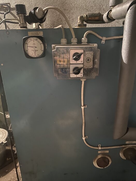 Gammal blå panna med tryckmätare och reglage i en källare, omgiven av rör och kablar.
