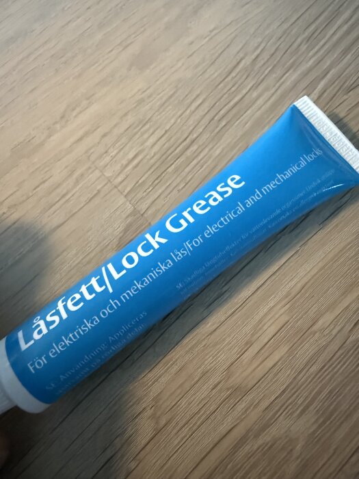 Blå tub med låsfett för elektriska och mekaniska lås ligger på ett träbord, texten på tuben är synlig.
