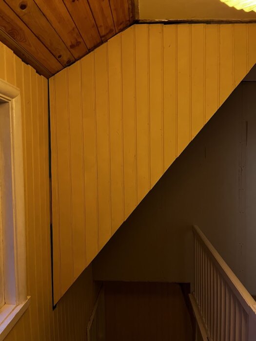 Vägg och snedtak med pärlspont monterat, synligt från ett trappräcke. Kombination av vinklar och träpanel på snedtaket. Fönster till vänster.