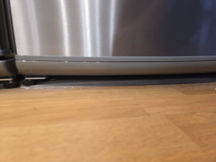 Bild på nederkant av kylskåpsdörr mot golvet, där dörrens gångjärn och otydlig bult syns.