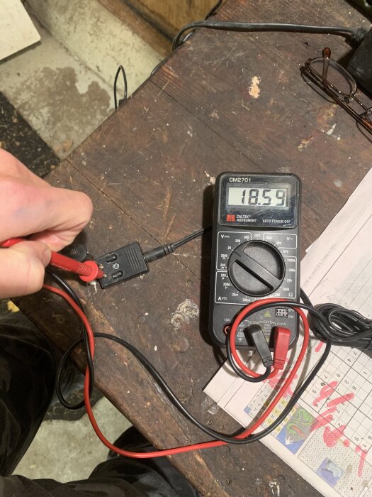 Digital multimeter visar 18,59 volt medan en person mäter spänningen på en laddare på ett träbord.