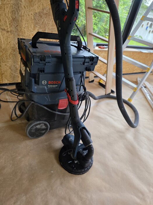 Bosch-maskin med dammsugare och slipmaskin i rum under renovering, med stege och träpanel i bakgrunden.