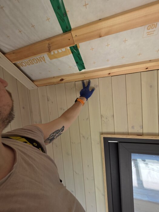 Person förbereder takfack för skorsten i rum med innertak i träpanel och synliga bjälkar.