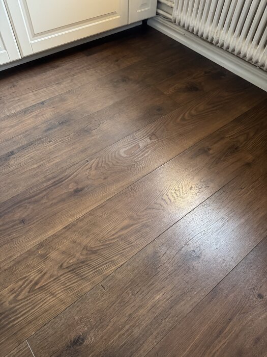Mörkt träparkettgolv med synlig ådring, installerat i kök nära vit köksskåpsfront och radiator.