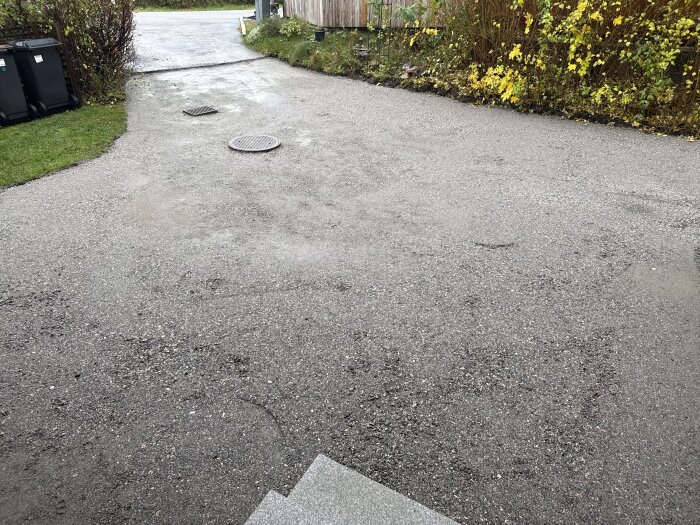 Uppfart där gammal asfalt tagits bort och marknivåer justerats, med en brunn och kant med gräs och buskar runtom.