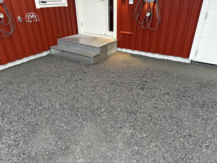 Nylagd grusyta framför röd byggnad utan asfalt, med justerade höjder, bredvid en entrédörr med trappsteg.