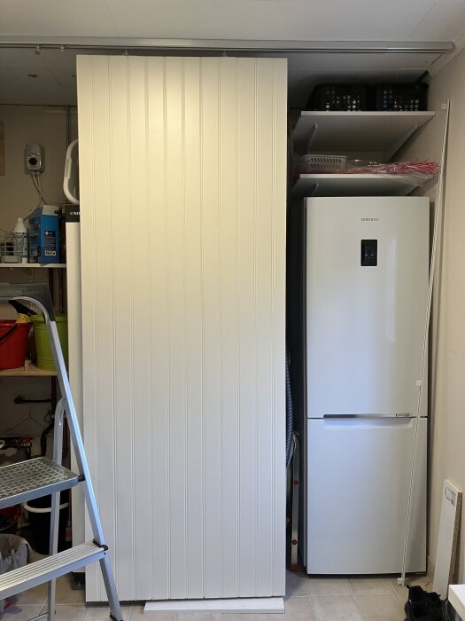 Vit dörr installerad bredvid ett kylskåp, med hyllor med förvaring ovanpå. Stege står bredvid. Döljer del av förvaringsutrymme.