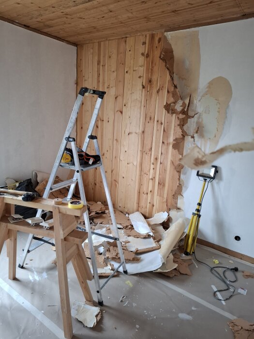 Rivningsarbete av väggpanel med stege, träplankor och verktyg i ett rum. Papper och dammigt golv indikerar pågående renovering.