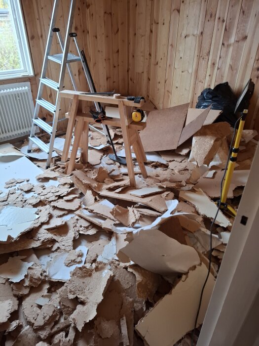 Rivningsarbete i trärum med gipsbitar på golvet, stege och verktyg synliga.