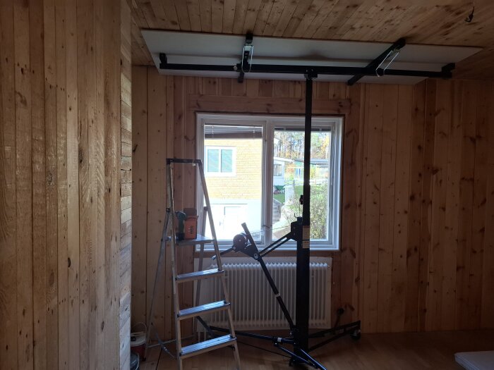 Rum med träväggar och verktyg, en stege och gipsskiva mot taket, radiator under fönster. Förberedelser för renovering.