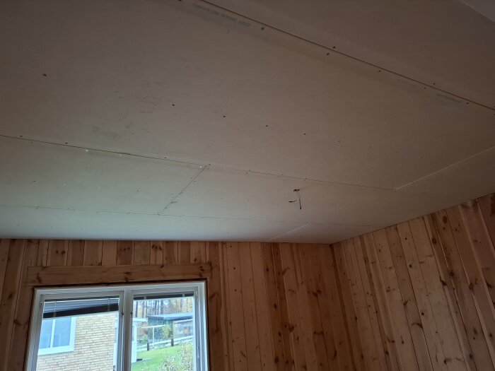 Nytt gipstak under installation i ett rum med träpanelväggar och fönster som visar utsikt mot grönska utanför.