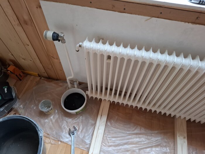Radiator demonteras med skiftnyckel, svart vatten i hink. Golvet skyddas med plast under renovering.