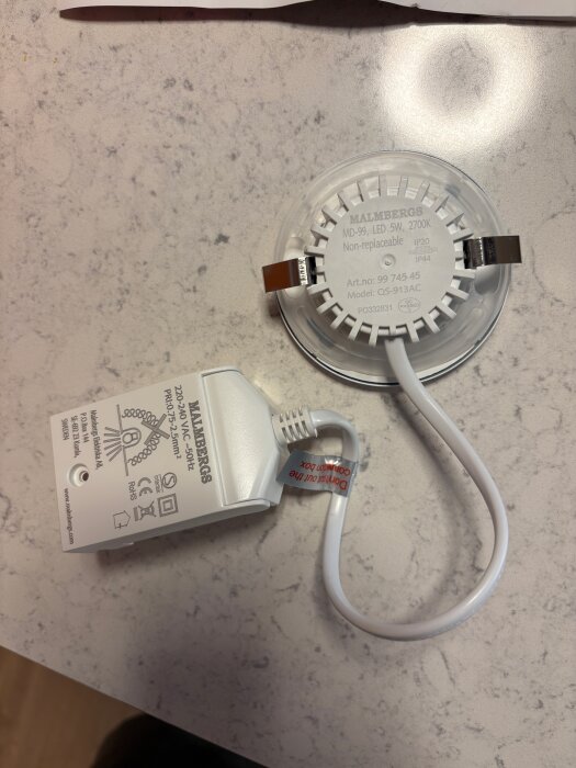 Malmbergs MD-99 LED-spotlight och driver med vit kabel och etikett på bord; etikett säger att kabeln inte får kapas.