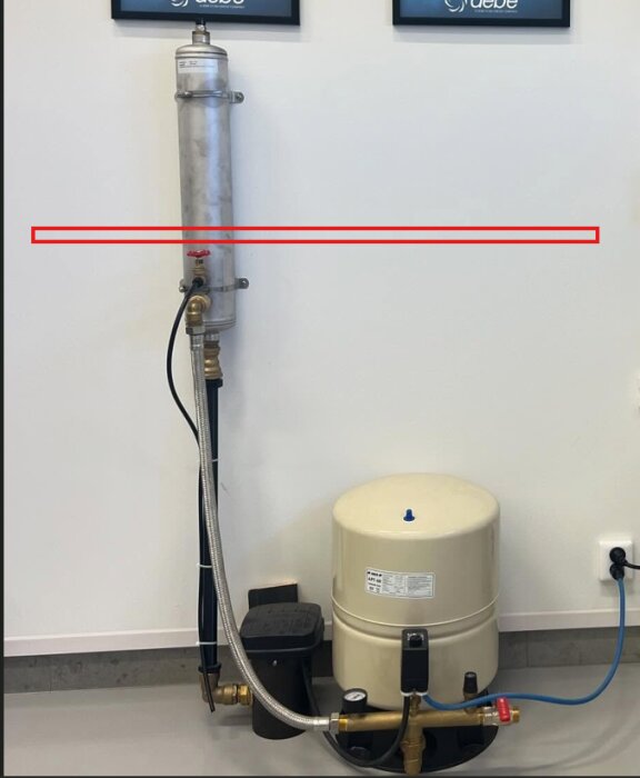 Syreaktivator monterad på väggen med röranslutningar och hydropress i förgrunden; röd markering och pil visar inkommande vatten.