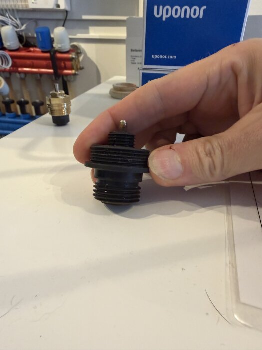 En hand håller i en svart ventil från en golvvärmefördelare; bakgrunden visar en Uponor-förpackning.