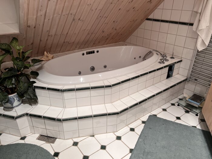 Inkastat badkar i ett badrum med lutande träpanelstak och omgivningar kaklade med vita plattor. Grön växt placerad vid badkarskanten.