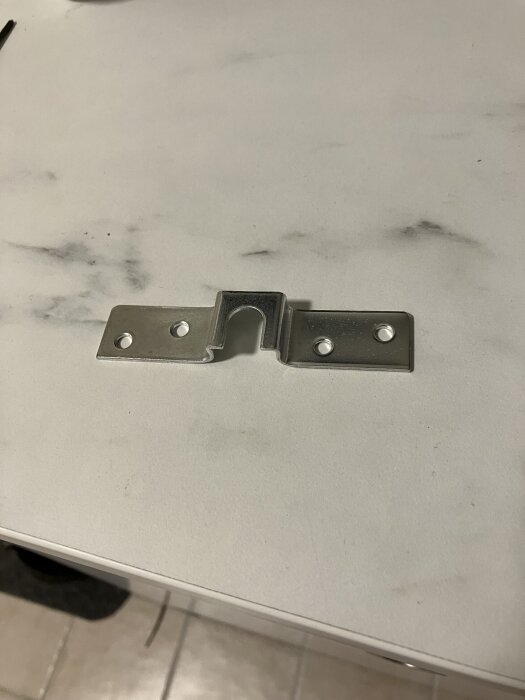 Metallfäste med osymmetrisk hålbild, visat på bord. Fyra hål, varav två är nyborrade för ökad hållfasthet, enligt inläggsbeskrivningen.