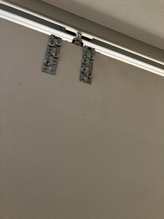 Två metalliska hålband med skruvar på en spånskiva för förstärkning, ovanför ett fäste med osymmetrisk hålbild.