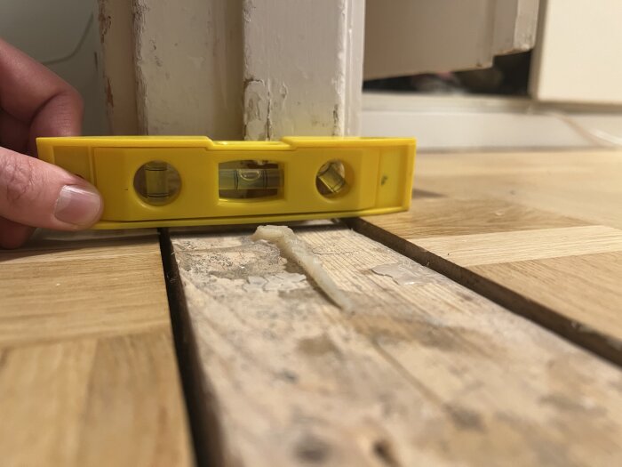 Gult vattenpass som visar nivåskillnad mellan två rumsgolv av trä vid en tröskel. Hand håller vattenpasset nära tröskeln.