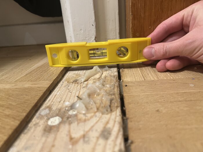 Golv och tröskel med nivåskillnad mäts med ett gult vattenpass, smuts och ojämnheter syns i skarven mellan två golvytor.
