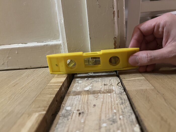 Gul vattenpass på tröskel mellan två sneda golv, en hand håller passet. Golven har en nivåskillnad på upp till 1 cm.