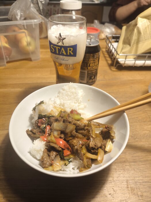 Risskål med stark mat och grönsaker, serverad med en öl i ett glas på ett träbord.