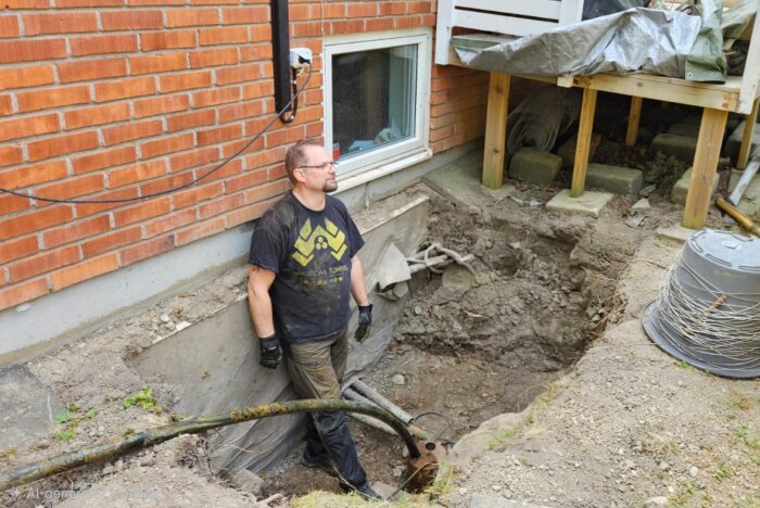 Person står i en grop intill husets fasad och dränering. Gropen är vid borrhålets anslutning för grundvattenpump, omgiven av lerjord.
