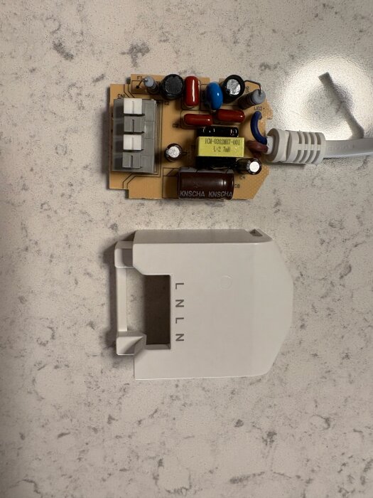 Kretskort till kopplingsbox med flera komponenter och en vit kabel; locket med märkning L N L N syns delvis bredvid.