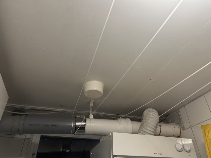 Innertak i tvättstuga med vitmålade skivor, ventilationsrör i plast och metall, samt vit tvättmaskin eller torktumlare med vridknappar.