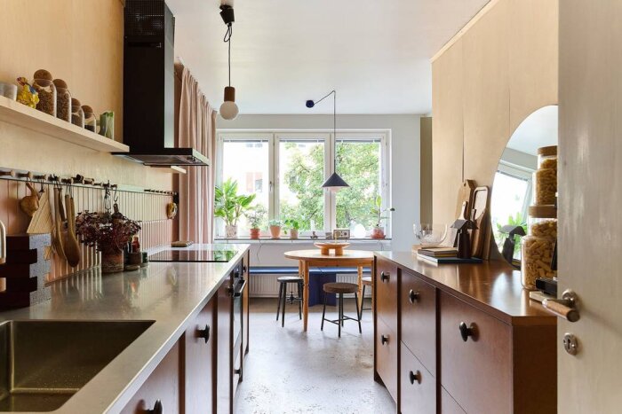 Ett modernt kök med MDF-skåp, stora fönster, rund spegel, träbord och växter i fönstret. Hängande lampor över bordet och inredning i neutral stil.