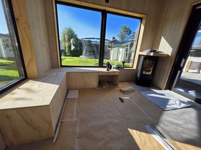 Platsbyggd sittmöbel av interiörplywood vid stora fönster, med vedförvaring och kamin i bakgrunden.