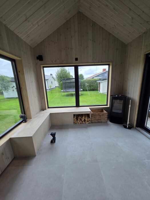 Platsbyggd sittmöbel under fönster med vedförvaring, betongliknande klinker på golvet, skruvdragare på sätet, kamin i hörnet, utsikt över trädgård.