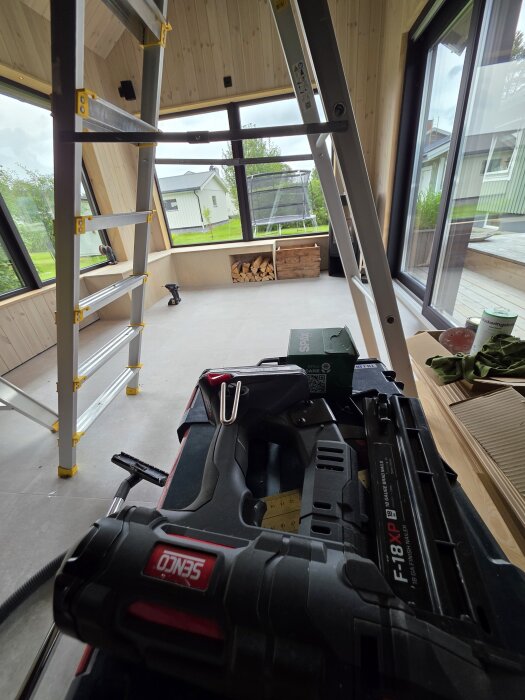 Interiörbild av ett bygge, med stege, plywoodväggar, och arbetsverktyg på golvet. Synligt utanför fönstret är grönska och närliggande hus.