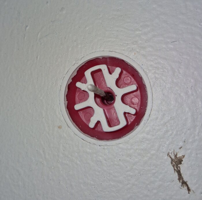 Beslag mitt i taket på balkong, nybyggd fastighet. Röd och vit form med metallstång, troligen för infästning eller upphängning.