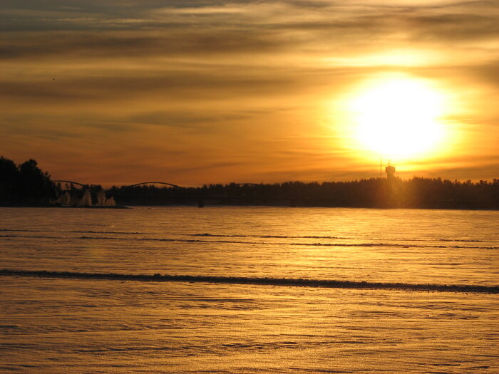 Solnedgång över fruset landskap i Luleå, 2007, med en bro och byggnader synliga i silhuett mot den gyllene himlen.
