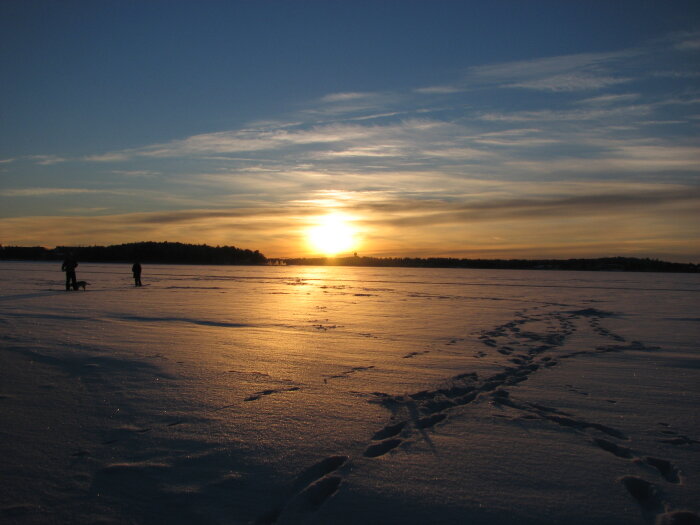 Solnedgång över snötäckt landskap i Luleå 2007 med fotspår i snön och personer i horisonten.