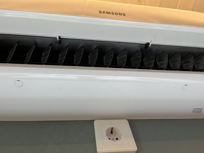 Samsung luftvärmepump med öppen framsida och synliga fläktblad, fotograferad ovanför ett eluttag.