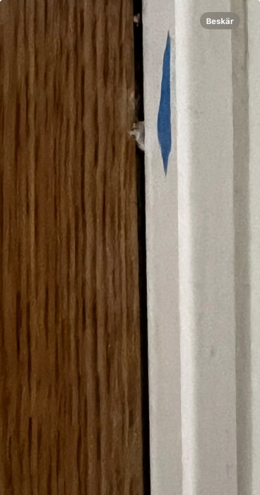 Glipa mellan parkettgolv och dörrkarm, cirka 1 cm bred, markerad med blå tejp mot en vit sockel.