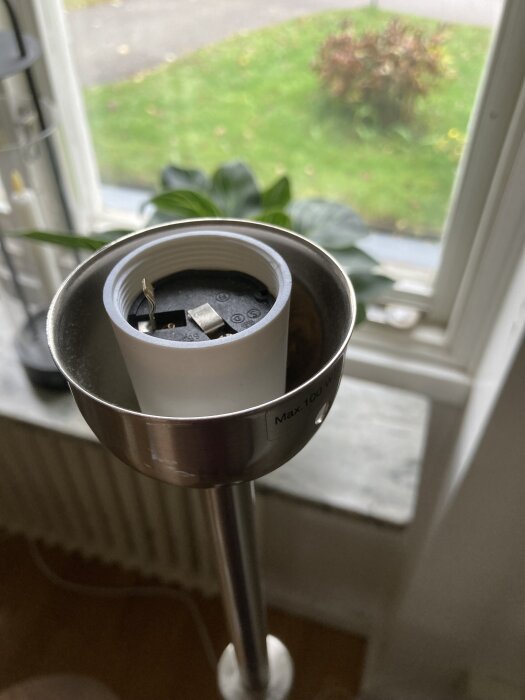 Öppen lamphållare på en golvlampa utan glödlampa, med dragsnöret synligt. Bakgrunden visar ett grönt fönsterlandskap.