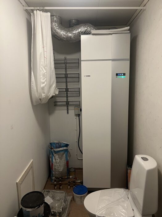 Rum med varmvattenberedare, toalettstol, och diverse byggmaterial; draperi hänger framför beredaren, fundering på gästtoalett.