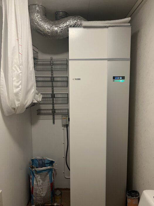 Rum med värmepump, hyllor och säckar med material. Draperi till vänster och vita väggar.