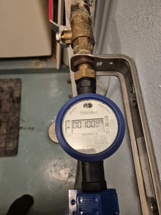 Digital vattenmätare med blå ram visar en förbrukning på 100,529 kubikmeter i en källare, med pilar för vattenflödesriktning.