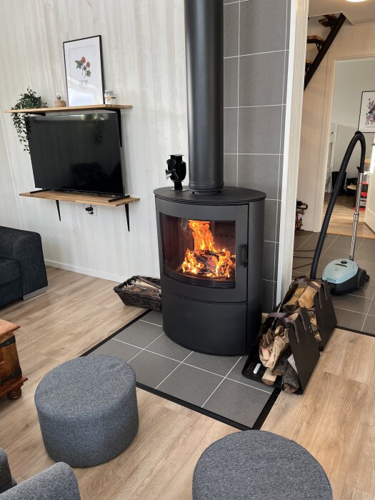 Svart vedeldad kamin med brasa i ett modernt vardagsrum, omgiven av trä och stiliga möbler; ved och dammsugare syns bredvid.