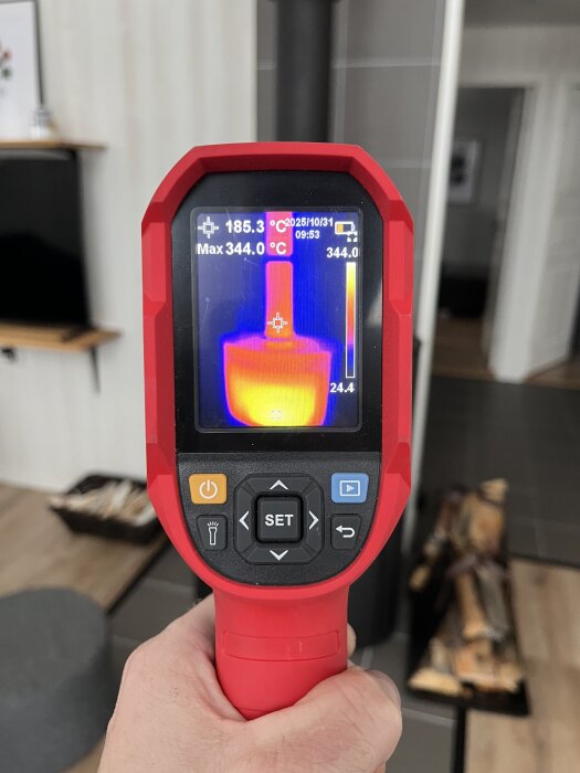 Hand som håller en värmekamera framför en kamin, visar temperaturer på 185,3°C och max 344,0°C. Bakgrund med ved och modern inredning.